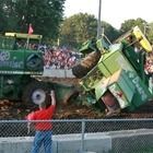 Upside-down green truck in a muddy field with a crowd behind a fence; person in red raises an arm in front.