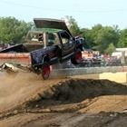 Pickup truck launching off a dirt ramp at an off-road track, with dust flying and spectators nearby
