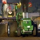 Green tractor at night in a lit dirt arena, towing a heavy sled with large rear tires.