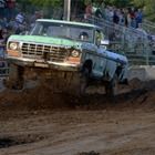 Blue pickup truck racing through muddy dirt track, splashing mud at an off-road event