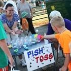 People playing a fish toss game at a fair booth with a sign reading “FISH TOSS”