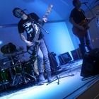 Live band performing on a blue-lit stage, with a singer at center, drummer left, and guitarist right.