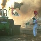 Person in white waving a red flag near a smoky tractor in dusty outdoor scene.