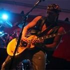 Guitarist in a hat playing an electric guitar on a dimly lit stage with blue and red lights.
