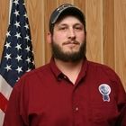 Man in red shirt and cap standing before wood paneling with an American flag behind him