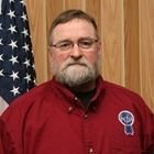Man in a red shirt standing before a wood-paneled wall and U.S. flag
