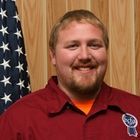 Smiling man in a red shirt standing before an American flag and wood-paneled wall