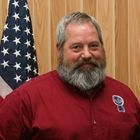 Man in red shirt smiling in front of a wood-paneled wall with an American flag.