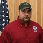 Man in a red button-up shirt and green cap stands indoors beside a U.S. flag.