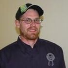 Man in glasses and green cap wearing a dark shirt, standing indoors against a plain wall.
