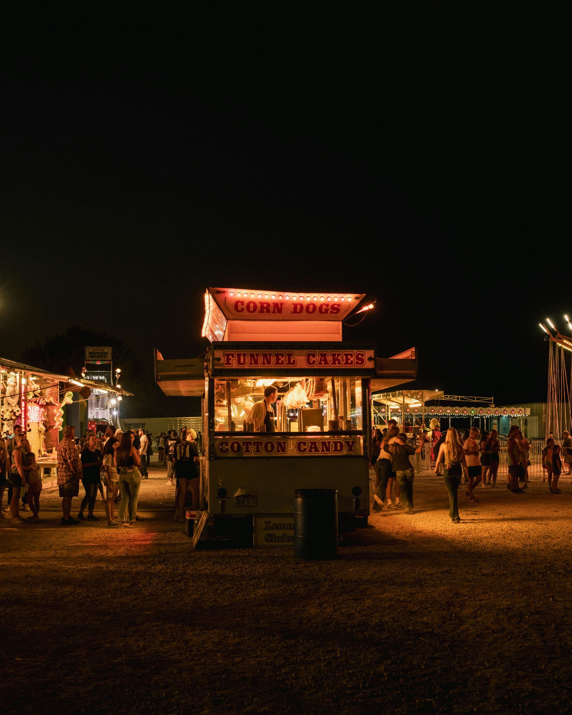 Nighttime outdoor festival with a lit food stall and people walking around on a dark gravel field.