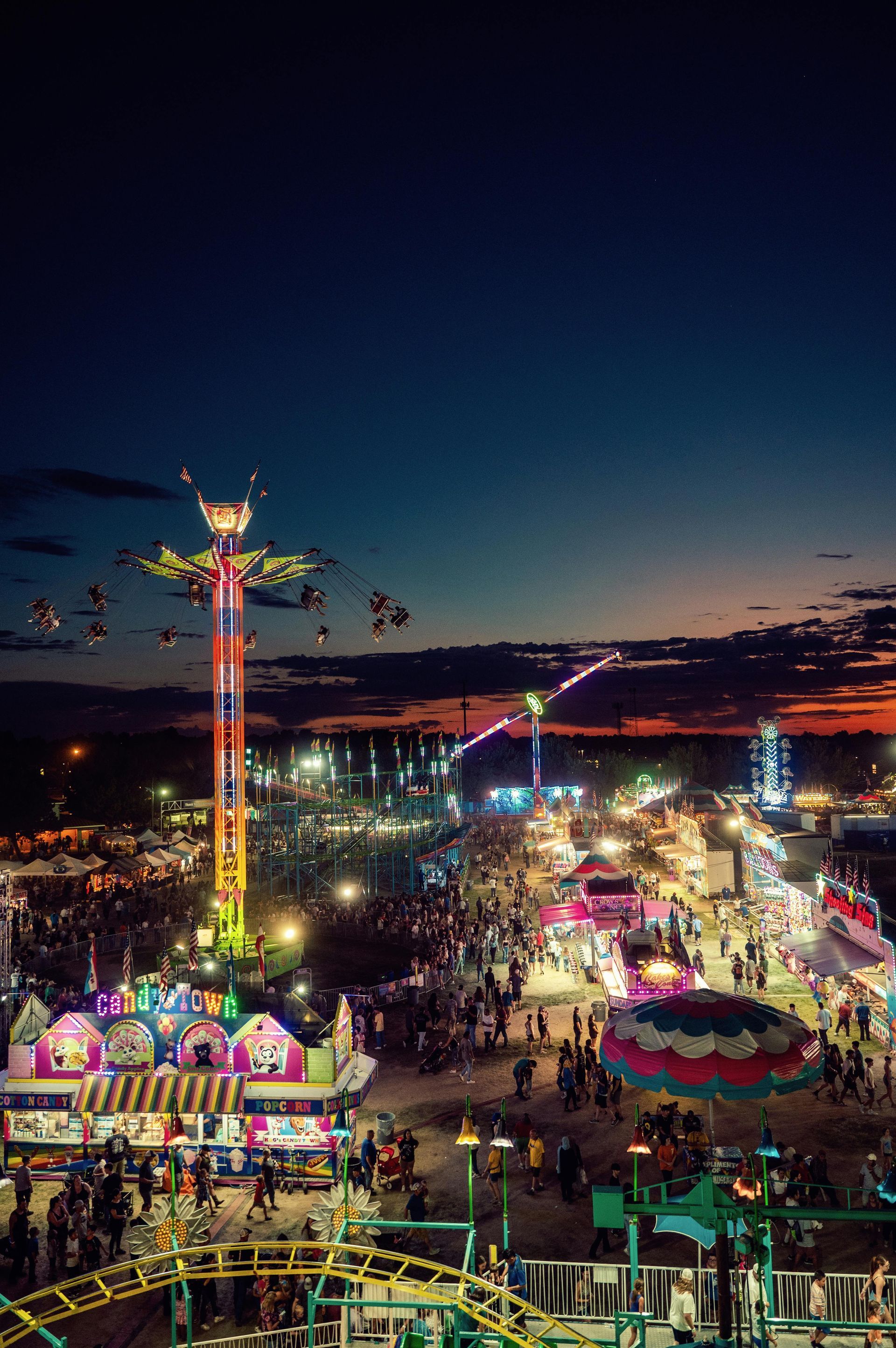 Nighttime carnival with colorful rides, bright lights, and a tall illuminated tower against a dark sky