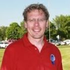 Smiling person in a red polo shirt standing outdoors near parked cars and trees