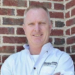 Man with short hair smiles, arms crossed, wearing white shirt with logo against brick wall.