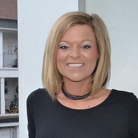 Woman with blonde hair, smiling, wearing a black shirt and necklace, standing outdoors.