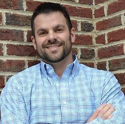Man with beard in blue plaid shirt, smiling in front of a brick wall.