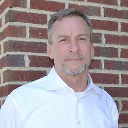 Man in white shirt, light beard, standing by a brick wall.