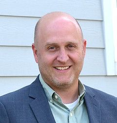 Bald man, smiling, wearing a blue blazer and green shirt, in front of a light blue wall.