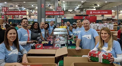 Volunteers packing gift boxes at an event. People wear blue shirts, sorting items in a large warehouse.