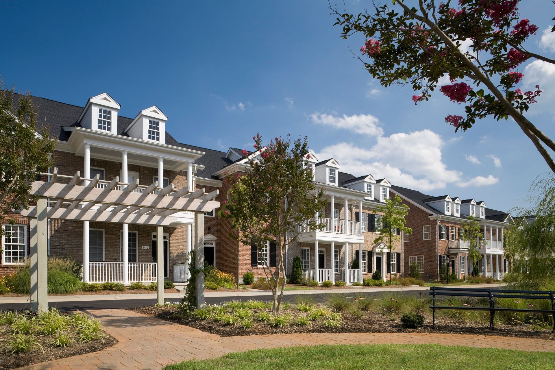 Row of brick townhouses with white balconies, under a blue sky, with trees in the foreground.