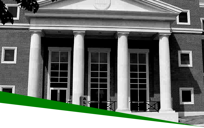 Black-and-white photo of a brick building with white columns and a green diagonal banner at the bottom