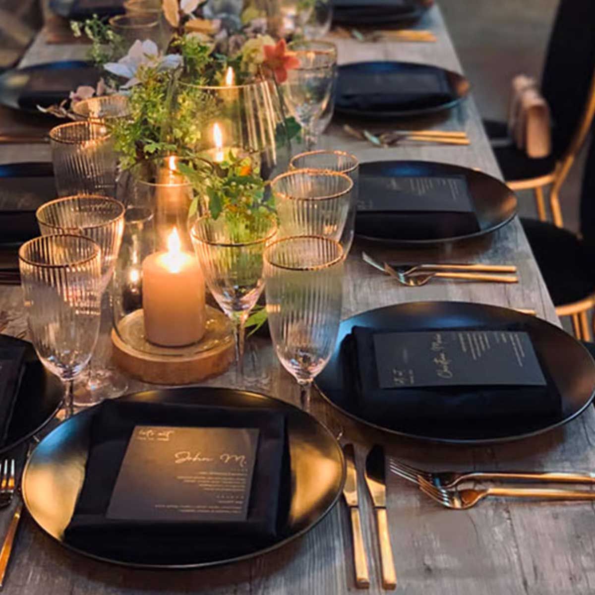 A beautiful gold and black table scape of plates , glasses , candles , and napkins set up for a private chef dinnner