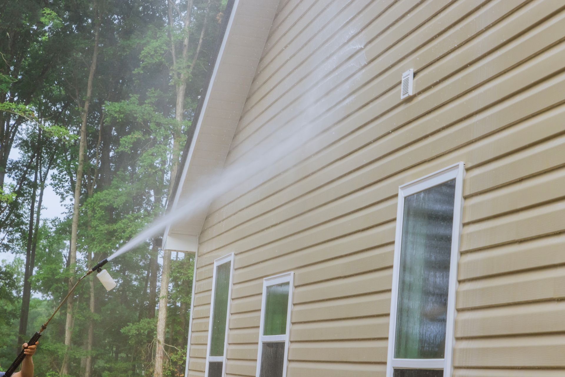 Person pressure washing tan vinyl siding on a house with white trim.