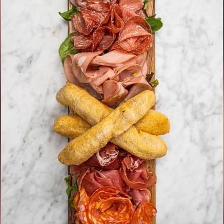 Un tagliere di salumi con affettati piegati, salame e due bastoncini di pasta fritta incrociati su una superficie di marmo bianco.