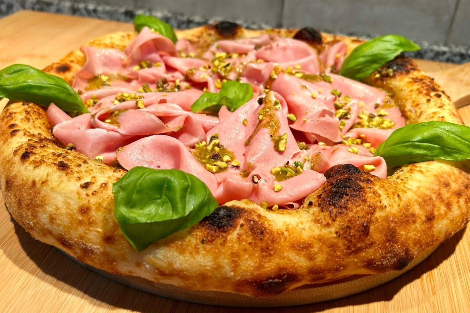 Una pizza cotta nel forno a legna, condita con mortadella tagliata a fettine sottili, pistacchi tritati e foglie di basilico fresco, servita su un tagliere di legno.