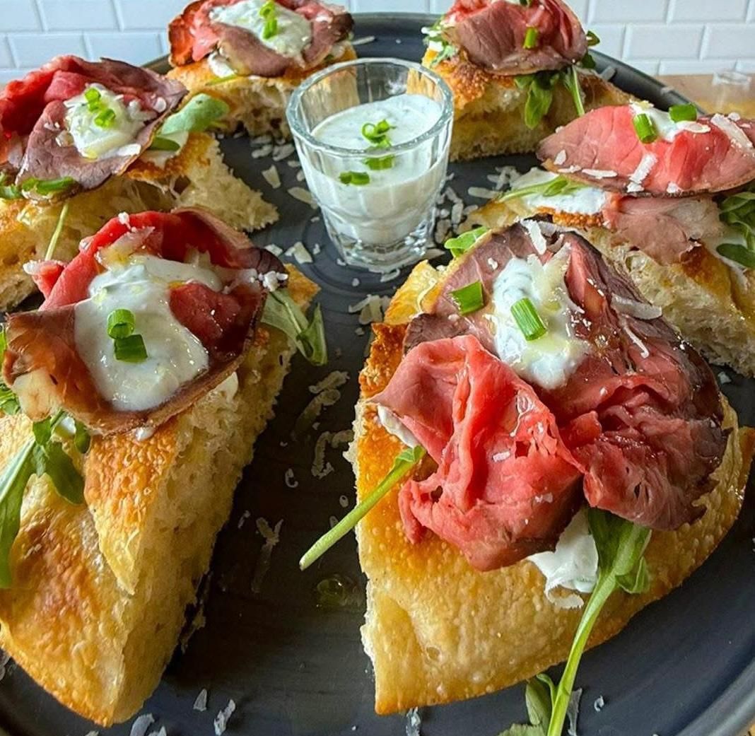 Diverse fette di focaccia farcite con roast beef, rucola e salsa bianca, servite con una ciotolina di salsa.