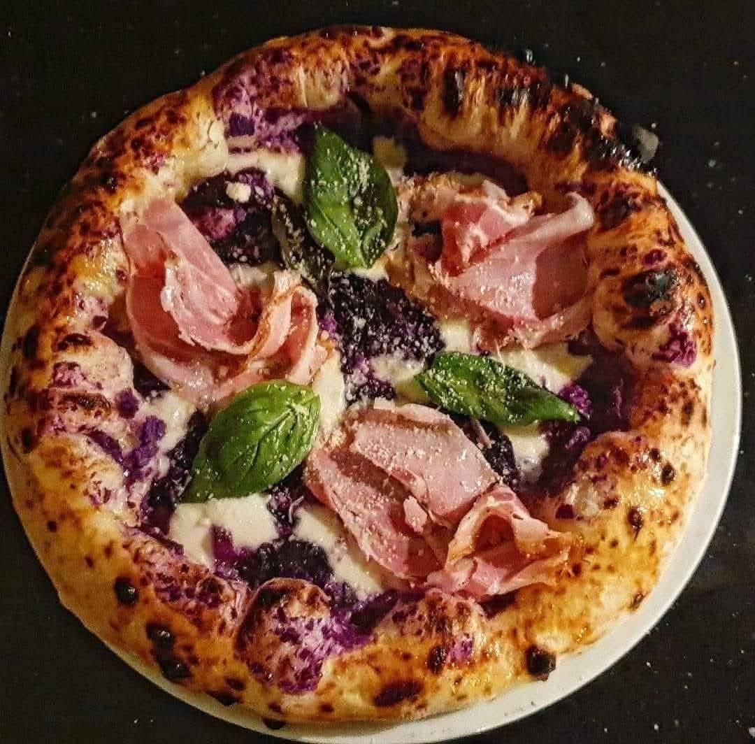 Una pizza cotta nel forno a legna con cavolo viola, formaggio fuso, fette di prosciutto ripiegate e basilico fresco su una base scura.