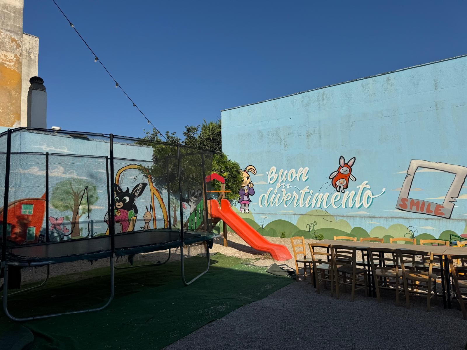 Un parco giochi all'aperto con un trampolino, uno scivolo e un muro blu dipinto con personaggi dei cartoni animati e scritte in italiano.