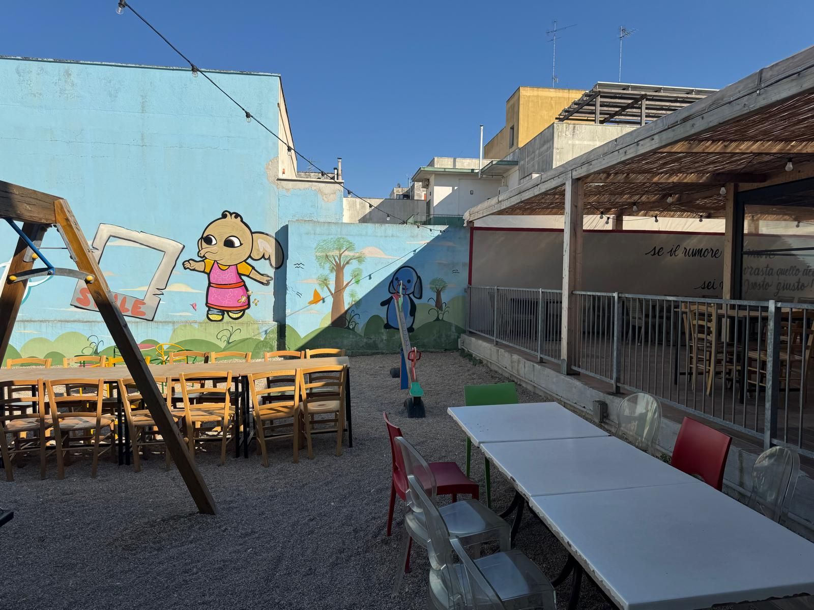 Patio esterno con pavimento in ghiaia, tavoli, sedie e un murale dipinto raffigurante un personaggio dei cartoni animati su una parete blu.