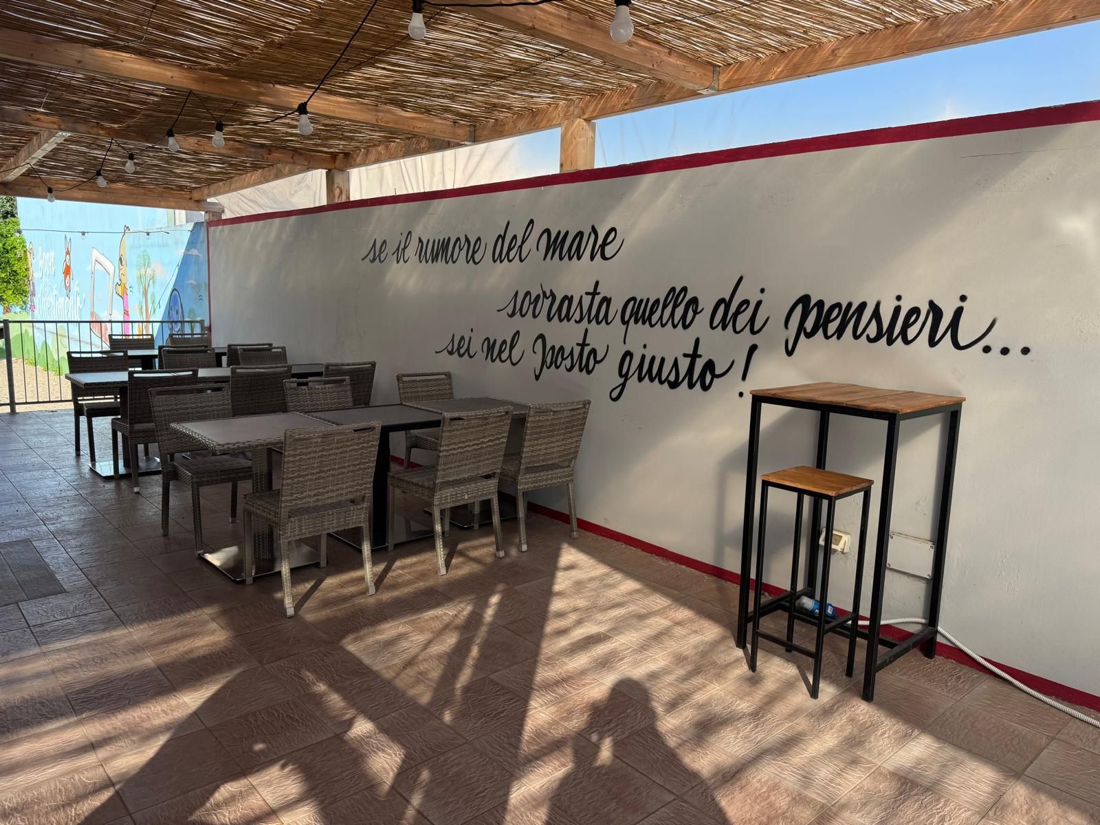 Terrazza esterna con tavoli e sedie, caratterizzata da una parete bianca con scritte in corsivo italiano sotto un tetto a doghe.