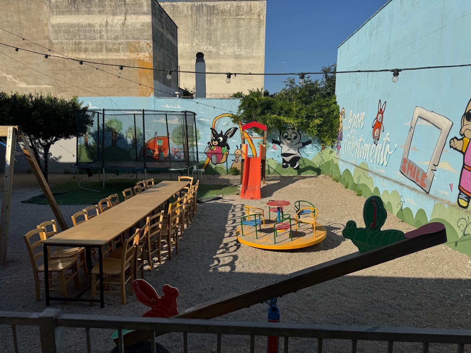 Un parco giochi all'aperto con un lungo tavolo da pranzo in legno, uno scivolo, una giostra, un trampolino elastico e un muro decorato con un murale.