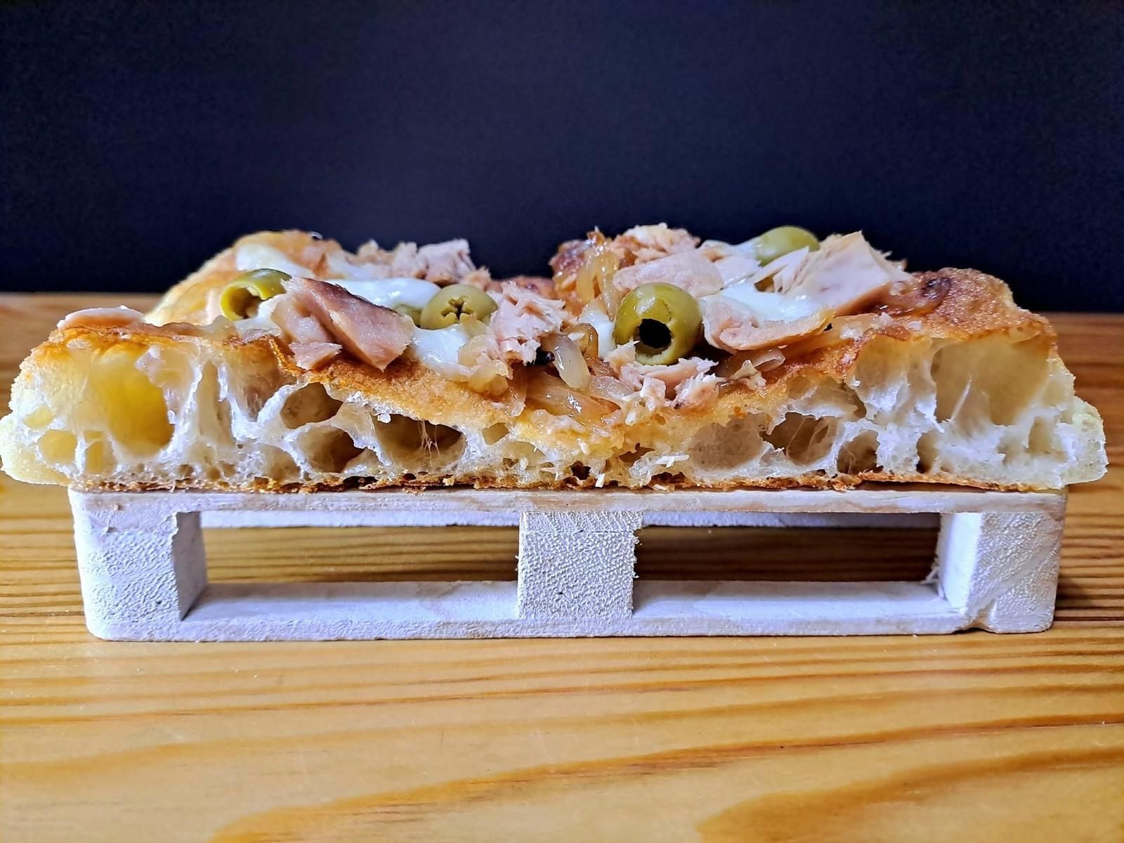 Una fetta di pizza con olive e prosciutto è adagiata su un piccolo vassoio di legno bianco, a sua volta appoggiato su una superficie di legno contro uno sfondo scuro.