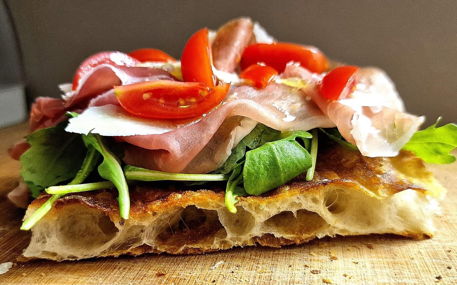 Una fetta di focaccia condita con rucola, prosciutto crudo, scaglie di parmigiano e pomodorini, servita su un tagliere di legno.