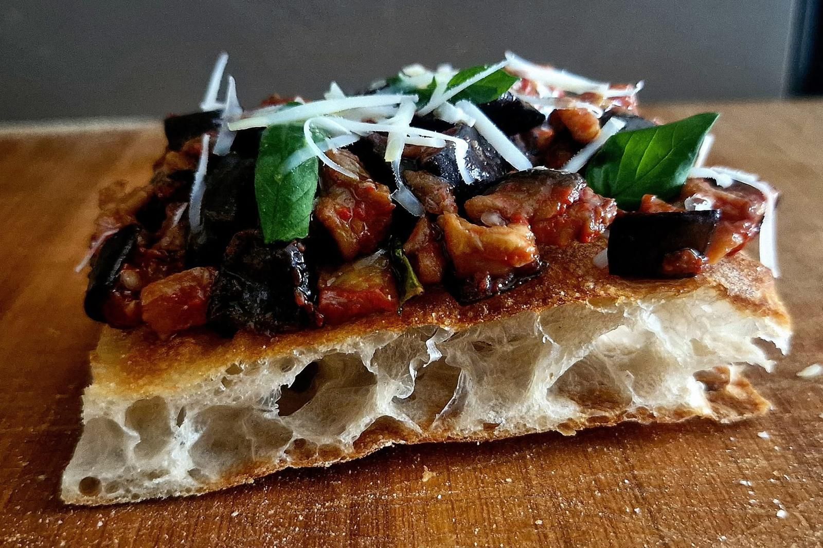 Una singola fetta quadrata di focaccia condita con melanzane saltate, pomodoro, basilico fresco e parmigiano grattugiato.
