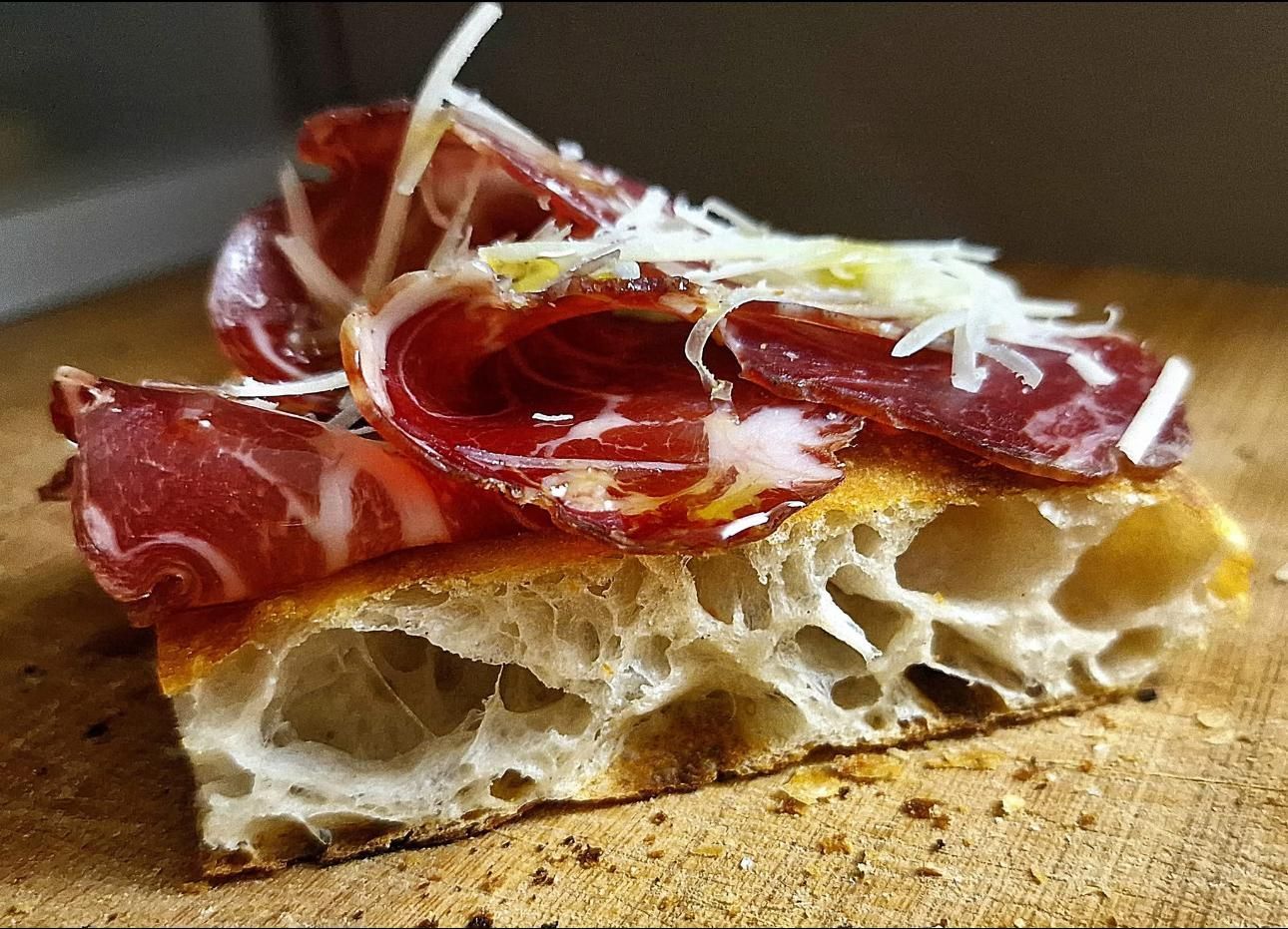 Una fetta ravvicinata di focaccia croccante e soffice, guarnita con sottili fette di salumi marmorizzati e formaggio bianco grattugiato.