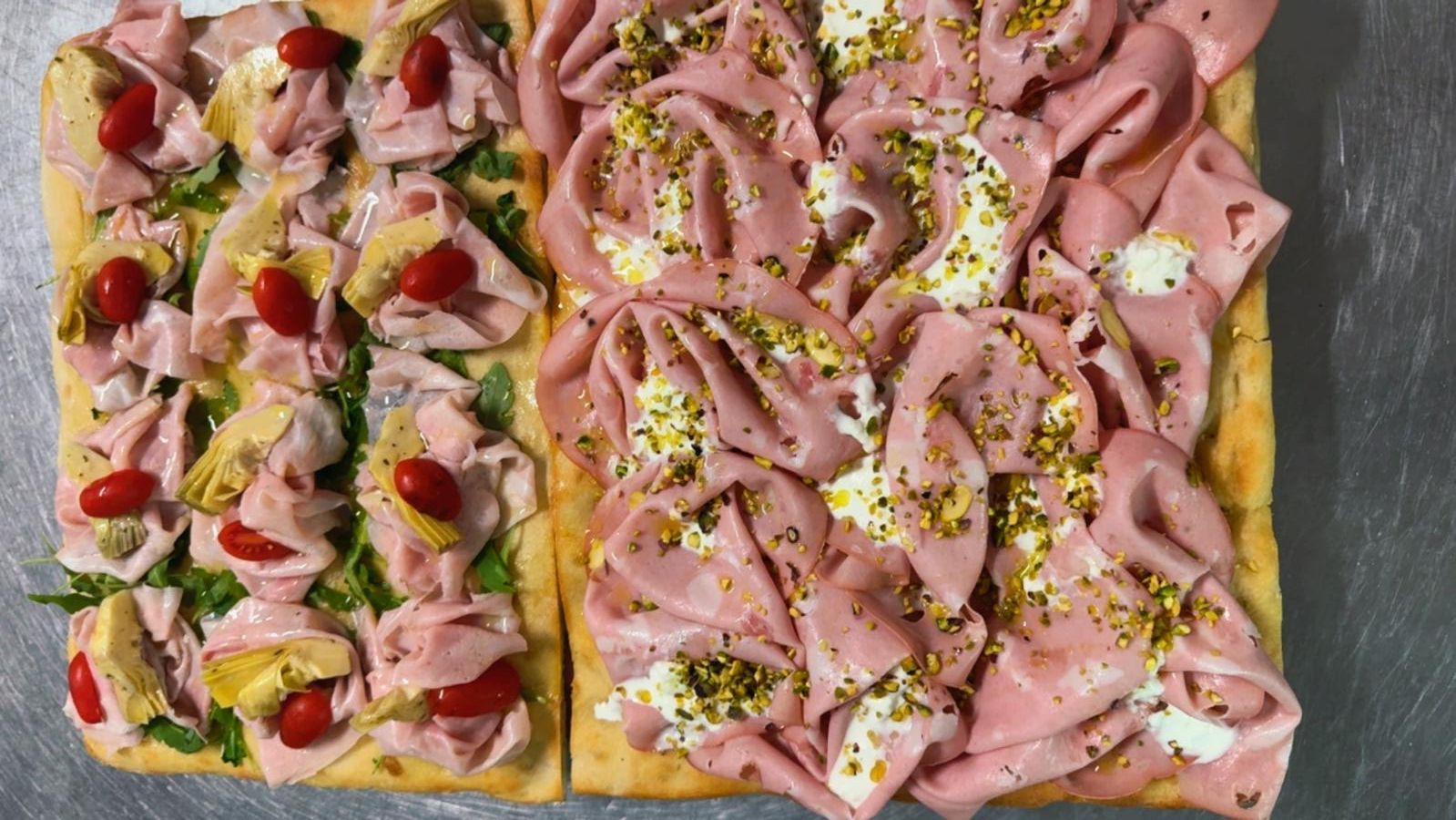 Due pizze rettangolari a base di focaccia, farcite con prosciutto a fette, carciofi, peperoni rossi e crema di formaggio con pistacchi.