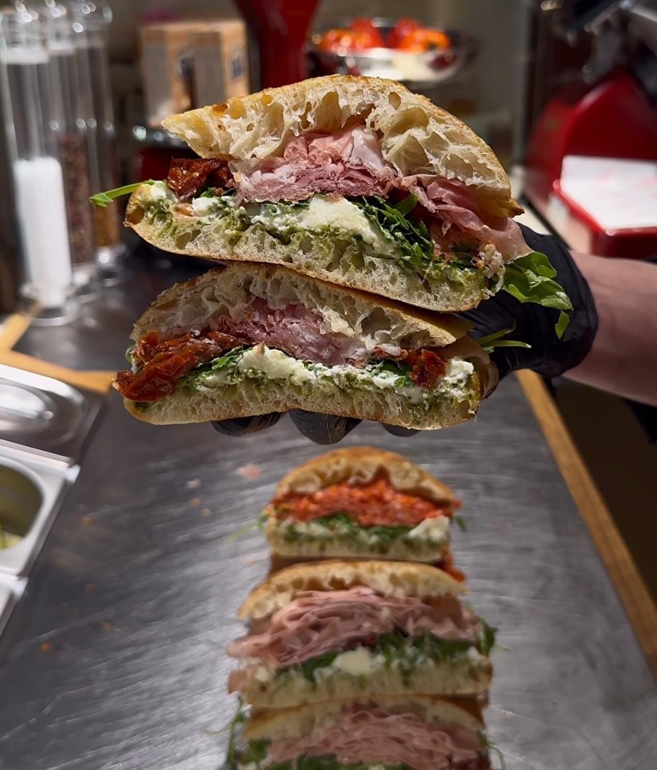 Una mano guantata regge un panino gourmet tagliato a metà, impilato sopra altri tre panini su un bancone metallico.