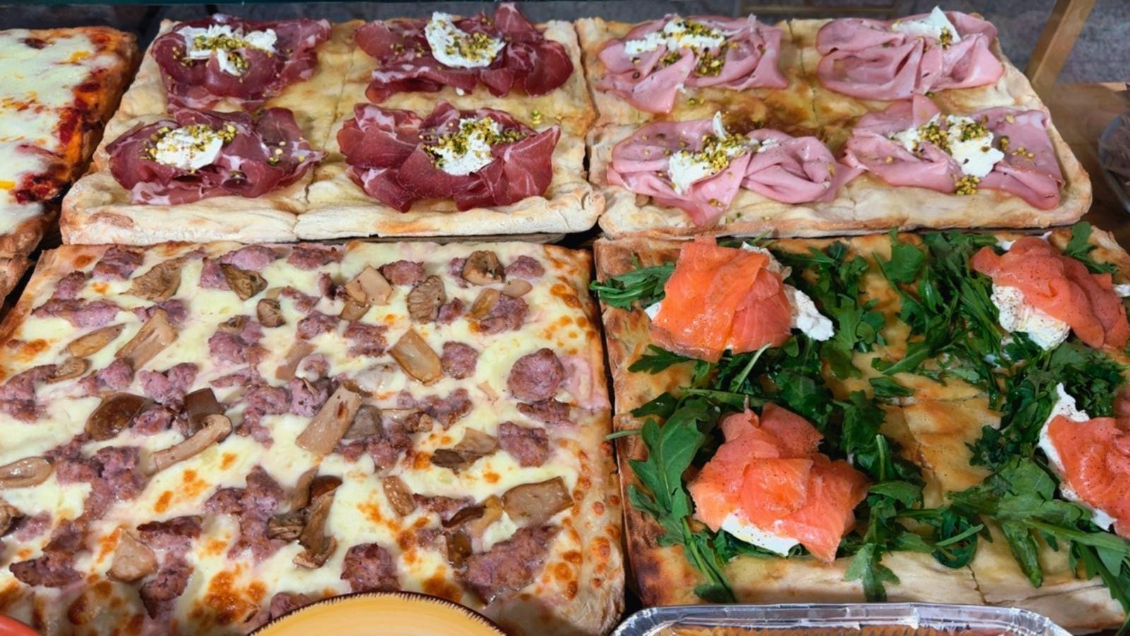 Quattro varietà di pizza rettangolare esposte in una vetrina da forno, con condimenti come prosciutto, mortadella e salmone.