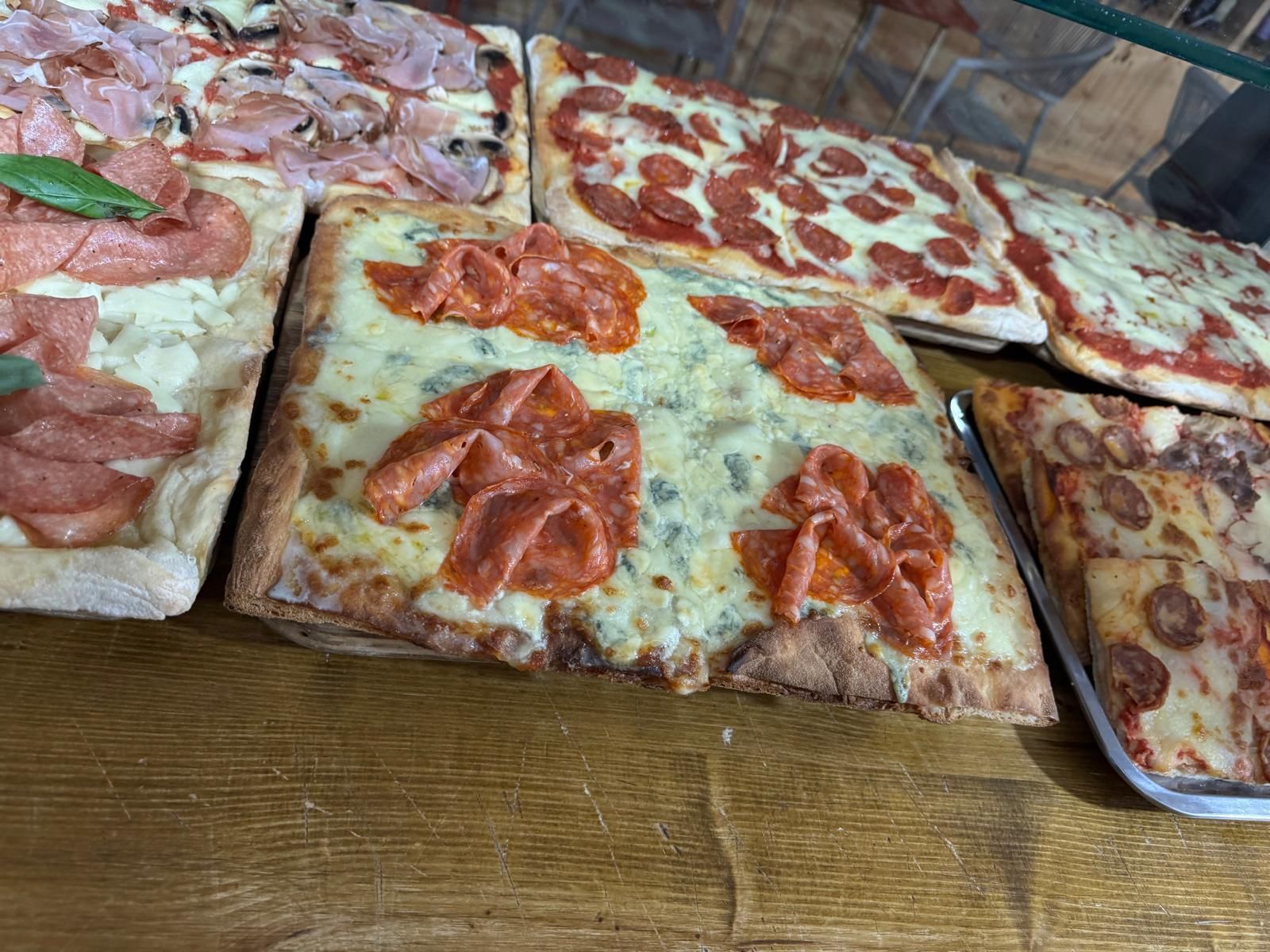 Una vetrina con varie pizze rettangolari con condimenti di peperoni, prosciutto e formaggio, disposte su un bancone di legno.