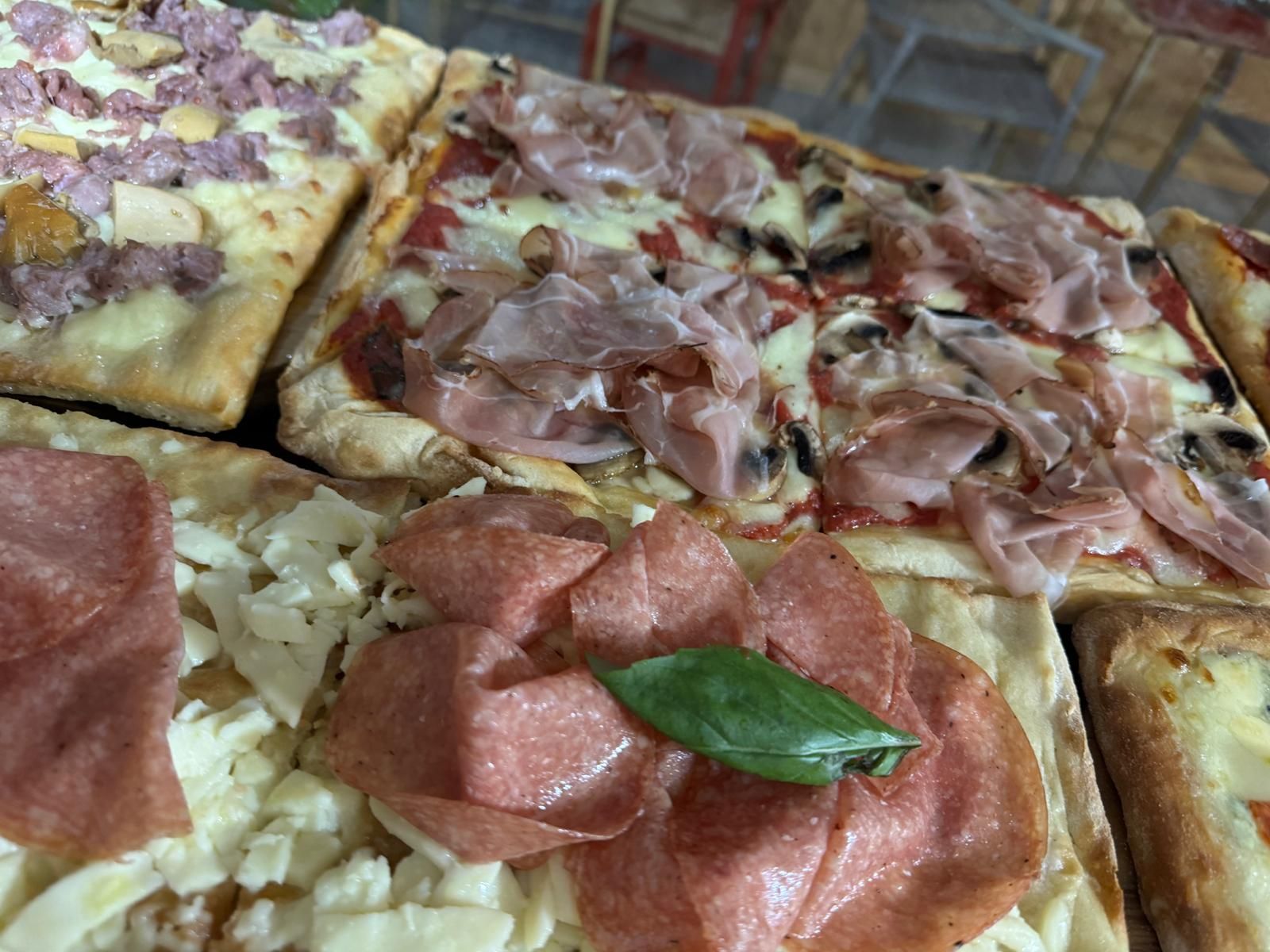 Un primo piano di diverse pizze rettangolari con vari condimenti, tra cui prosciutto cotto, prosciutto crudo, salame e basilico fresco.
