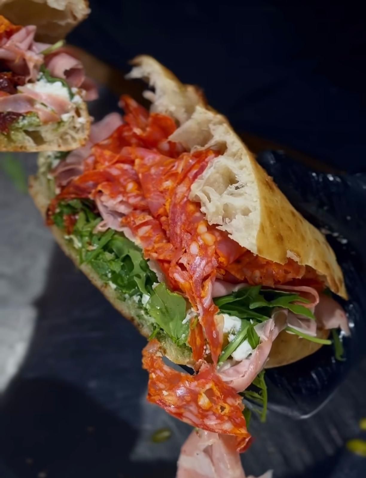 Una persona con i guanti neri tiene in mano un panino artigianale diviso a metà, farcito con salame tagliato finemente, prosciutto, verdure e formaggio.