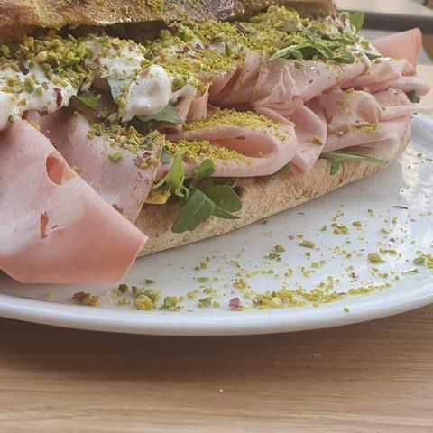 Un panino su un piatto bianco, con mortadella piegata, formaggio bianco cremoso e una generosa spolverata di pistacchi tritati.