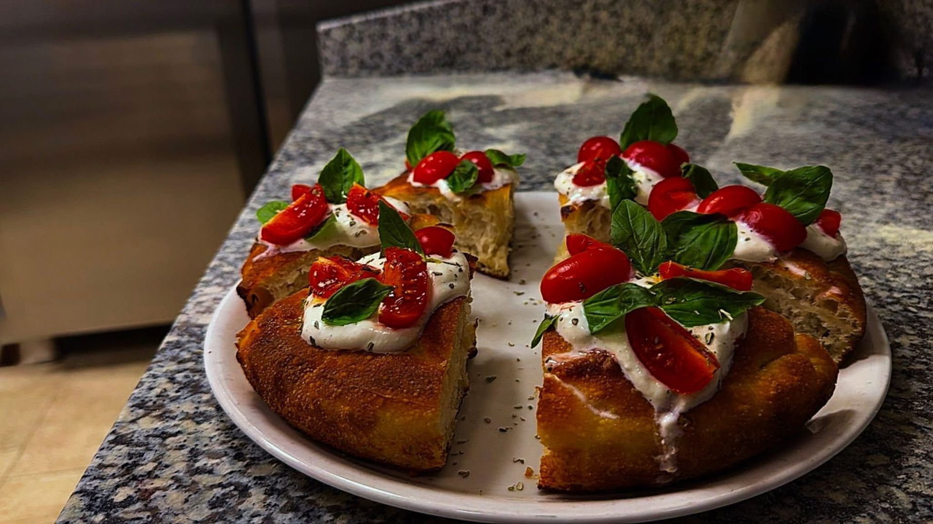 Un piatto di fette di pizza condite con formaggio bianco, pomodorini rossi e basilico fresco, adagiato su un piano di lavoro in granito.