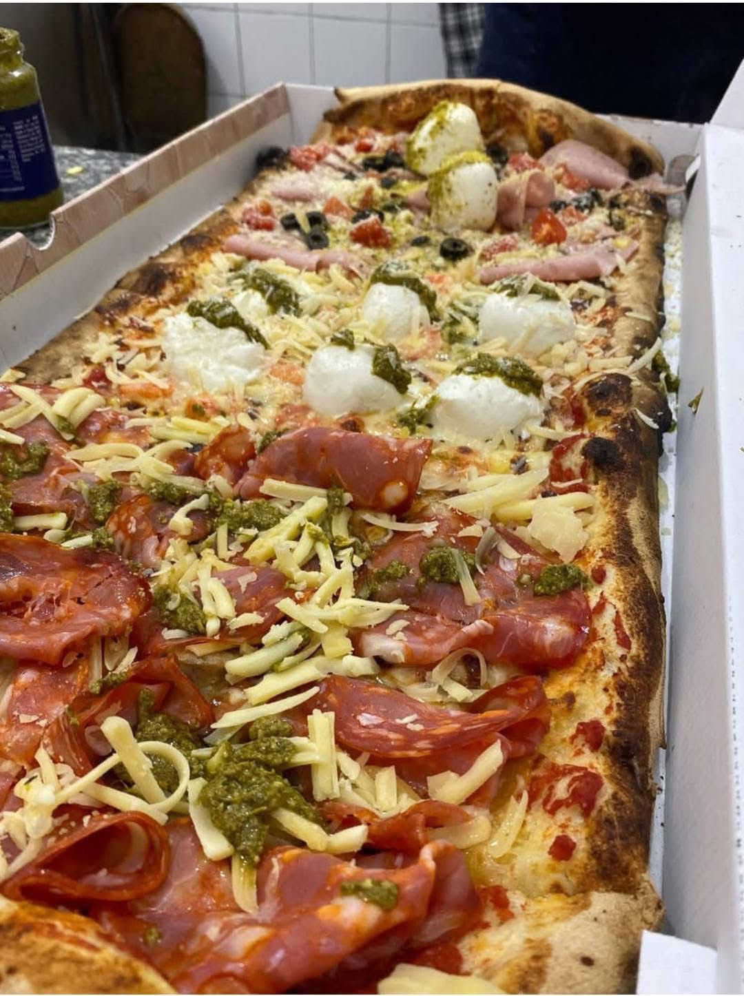 Una pizza lunga e rettangolare in una scatola di cartone, condita con salame, formaggio grattugiato, pesto e ciuffi di formaggio fresco.