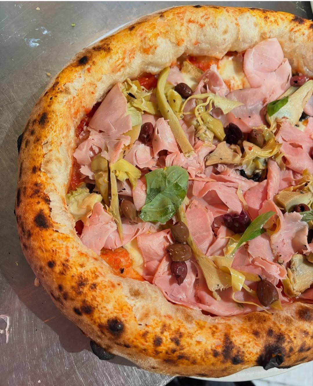 Una pizza cotta nel forno a legna, condita con sottili fette di prosciutto, carciofi, olive e basilico fresco su una base di pasta chiara.