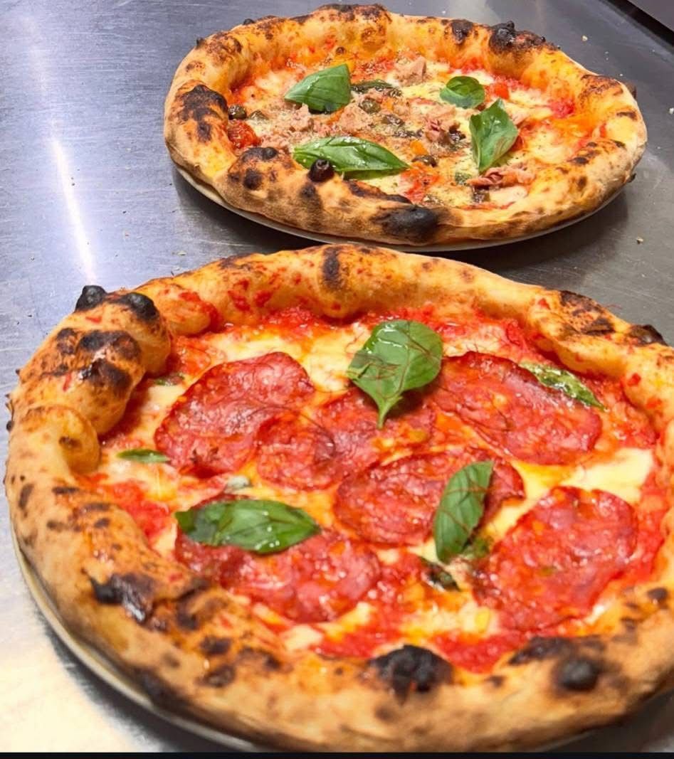 Due pizze cotte nel forno a legna, condite con salame, salsa di pomodoro, mozzarella e basilico fresco, adagiate su una superficie metallica.